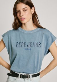 Mėlynas medvilninis trumpas rankovė t-shirt su "PEPE JEANS LONDON" tekstu džinsų įkvėpto siuvinėjimo stiliaus. Sujungtas su juodu diržu ir šviesiais džinsais.