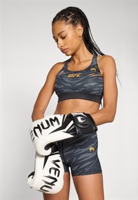 Top y pantalones cortos deportivos en negro y gris con un patrón de camuflaje. El modelo sostiene guantes de boxeo en blanco y negro con acentos de marca.