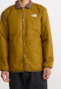 Mężczyzna w musztardowej żółtej kurtce North Face z brązowym kołnierzem i wykończeniem, stojący na tle jednolitej białej ściany.