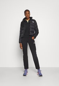 Piumino nero con zip frontale, pullover nero, pantaloni neri e scarpe da ginnastica grigio-viola, indossati da una persona in piedi contro uno sfondo bianco.
