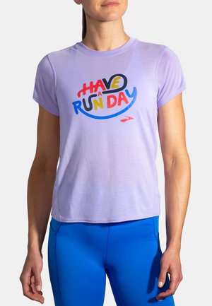 Kobieta ubrana w jasnofioletową koszulkę z krótkim rękawem z kolorowym napisem „Have a Run Day” oraz jasnoniebieskie legginsy, stojąca na białym tle.