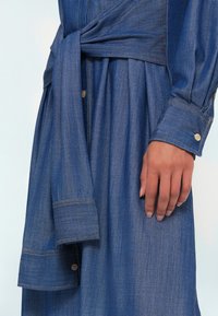 Γυναικείο φόρεμα από τζιν σε μέτριο μπλε. Διαθέτει ζώνη στη μέση, κουμπιά στα μανίκια και φαρδύ φουστάνι με λεπτή γυαλάδα και υφή.