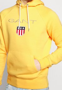 Žlutá mikina z bavlněné směsi, s klokankovou kapsou, kapucí se stahovací šňůrkou a vyšívaným štítem s motivem hvězd a pruhů.