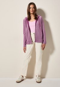 Haut à capuche zippé lilas avec cordon de serrage, associé à un débardeur rose, un pantalon droit crème, et des baskets blanches avec des accents bruns.