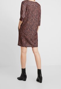 Nainen seisoo kädet taskuissa, päällään polveen ulottuva leopardinkuosinen mekko ja mustat nilkkurit, joissa on korot ja vetoketjut.