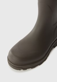 Botte en caoutchouc marron avec un bout arrondi et une semelle texturée, présentant une finition lisse et brillante ainsi que des éléments de design subtils sur l'empeigne.