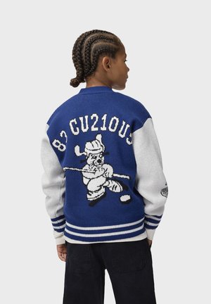Persona con capelli intrecciati indossa una giacca varsity blu e bianca con un giocatore di hockey dei cartoni animati e il testo "82 CU210US" sul retro.