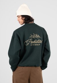 Uomo con un berretto lavorato a maglia bianco, felpa verde scuro con la scritta "Prohibited 10119 Berlin" e un design di foglia sul retro, e pantaloni marroni.