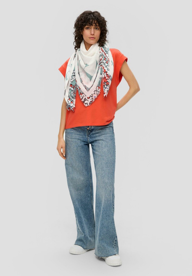 Oranje top met cap sleeves en een losse fit, gecombineerd met lichtblauwe flared jeans en een witte sjaal met een patroon van bloemdessins en franjes.