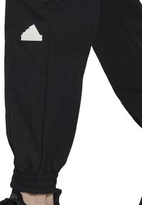 Svarta joggerbyxor i lätt tyg, med elastisk mudd och en vit gummimärke med Adidas-logotyp på vänster lår.