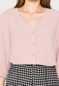 Blouse rose clair en V avec des manches courtes et quatre boutons sur le devant ; tissu texturé. Associée à une jupe pied-de-poule noir et blanc.