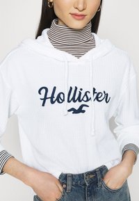Sweat à capuche blanc avec tissu texturé, arborant un logo "Hollister" bleu marine. Porté par-dessus un col roulé rayé noir et blanc. Bas en denim.