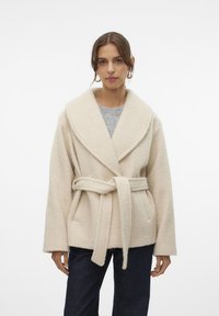 Beige fleecejacka med bred sjalkrag, bälte i midjan och synliga sömmar. Tyget har en mjuk textur; kombineras med mörka jeans.