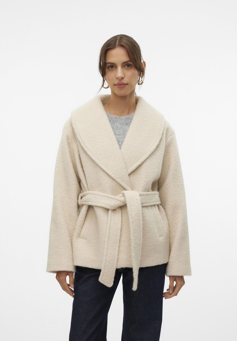 Beige fleecejacka med bred sjalkrag, bälte i midjan och synliga sömmar. Tyget har en mjuk textur; kombineras med mörka jeans.