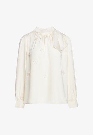 Blouse crème à manches longues avec col montant, petites appliques florales, tissu froncé près du col et détail nœud sur le côté.