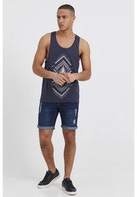 Mornarska majica bez rukava s bijelim geometrijskim uzorkom, uparena s tamnim traper kratkim hlačama i crnim tenisicama. Kratke hlače su postarane i imaju podvrnute rubove.