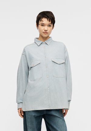 Mladá osoba s krátkymi tmavými vlasmi, oblečená v svetlomodrej pruhovanej košeli na gombíky a voľných tmavomodrých džínsoch, stojaca pred bielym pozadím.