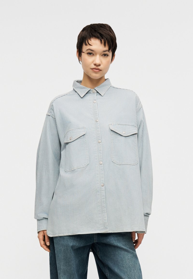 Νέος με κοντά σκούρα μαλλιά, φορώντας ένα ανοιχτό μπλε πουκάμισο με ρίγες και άνετο σκούρο μπλε τζιν, στέκεται μπροστά σε λευκό φόντο.