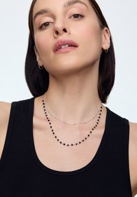 Naszyjniki warstwowe z małymi czarnymi koralikami na srebrnym łańcuszku, noszone na czarnej bluzce bez rękawów, z gładką, wypolerowaną powierzchnią.