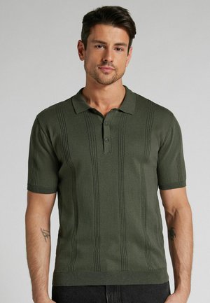 Mann trägt ein dunkelgrünes, kurzärmeliges Strick-Poloshirt mit vertikalen, strukturierten Streifen und steht vor einem schlichten grauen Hintergrund.