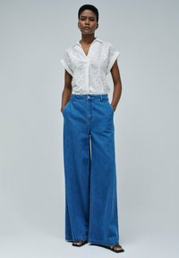 Blusa blanca de manga corta con bordado floral, combinada con jeans de mezclilla azul de pierna ancha y talle alto. El modelo está de pie con las manos en los bolsillos.