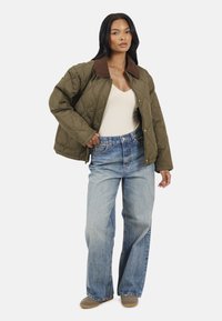 Veste matelassée vert olive avec un col marron, associée à un jean large bleu clair et des chaussures beiges. Design simple, look décontracté.