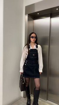 Femeie purtând ochelari de soare, cămașă albă, rochie de blugi neagră cu bretele, cizme negre până la genunchi și ținând o geantă maro, stând lângă ușile liftului.