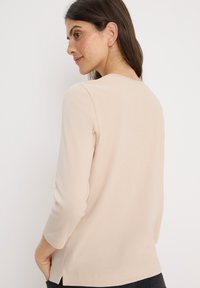 Beige ribbattu pitkähihaine paita, jossa on pyöreä kaula-aukko, sivuaukot ja istuva siluetti, valmistettu pehmeästä joustavasta kankaasta.