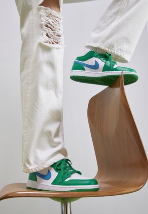 Jean blanc avec détail déchiré au niveau du genou, associé à des baskets vertes et blanches avec des accents bleus, placés sur une chaise en bois courbée.