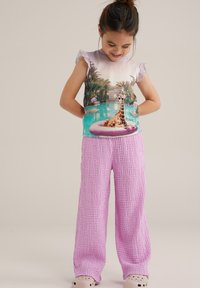 Majica s kratkimi rokavi z grafiko žirafe in palmami, v kombinaciji z teksturiranimi, ohlapnimi roza hlačami in bež crocsi.