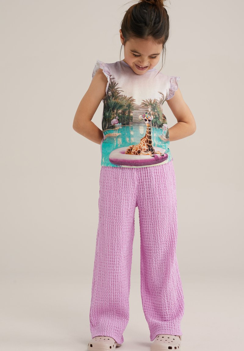 Majica s kratkimi rokavi z grafiko žirafe in palmami, v kombinaciji z teksturiranimi, ohlapnimi roza hlačami in bež crocsi.