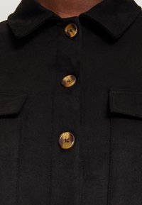 Svart skjorta med krage, prydd med tre bruna knappar och två bröstfickor med lock. Mjuk, strukturerad tyg.