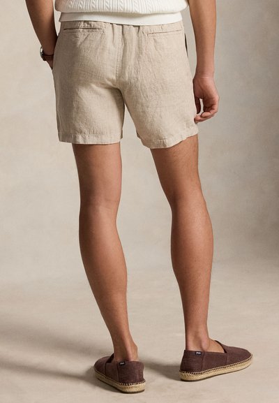Άνδρας που φοράει μπεζ λινά σορτς και καφέ espadrilles χωρίς κορδόνια, στέκεται με τα χέρια χαλαρά στο πλάι, μπροστά σε απλό φόντο.
