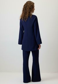 Completo blu navy con giacca su misura e pantaloni a zampa, caratterizzato da una texture liscia e un design semplice. La giacca ha maniche lunghe.