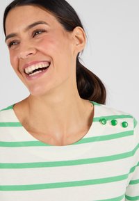Vrouw met donker haar in een staart die lacht, gekleed in een wit en groen gestreept shirt met drie groene knopen op de schouder.