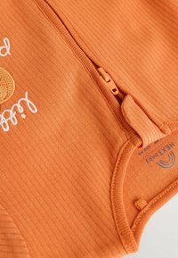Combinaison pour bébé orange à côtes avec fermeture éclair, ornée de texte brodé et d'une texture qui suggère la douceur. Logo imprimé à l'intérieur.