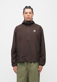 Καφέ ελαφρύ μπουφάν αδιάβροχο με κουκούλα, διαθέτει μπροστινό φερμουάρ και μικρό λευκό λογότυπο στο αριστερό στήθος. Υφασμένο ύφασμα με υφή.