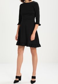 Robe noire à manches trois-quarts, ourlet à volants, corsage ajusté et tissu lisse. Associée à des sandales noires à talons blocs.