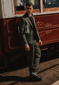 Žalias kostiumas su pritaikytu siluetu, dėvimas su juodu megztiniu. Juodi batai su storu padu. Stovi šalia raudono vintage tramvajaus.