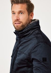 Dunkelblaue Jacke mit hohem Kragen, ausgestattet mit einem weichen grauen Futter, sichtbaren Nähten und einer mit Reißverschluss versehenen Tasche auf der linken Seite.