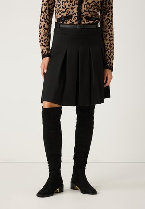 Schwarzer, plissierter Rock mit hohem Bund, kombiniert mit kniebedeckenden schwarzen Wildlederstiefeln mit goldenen Akzenten und einer Cardigan im Leopardenmuster.