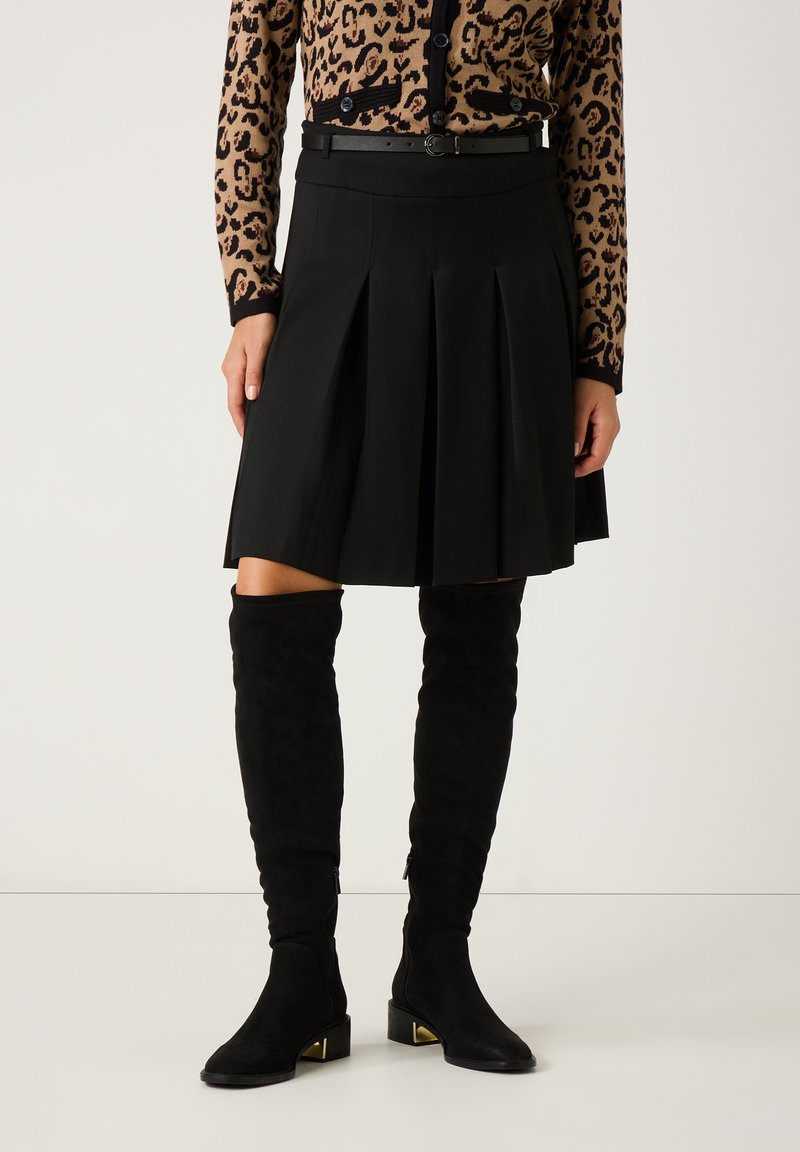 Schwarzer, plissierter Rock mit hohem Bund, kombiniert mit kniebedeckenden schwarzen Wildlederstiefeln mit goldenen Akzenten und einer Cardigan im Leopardenmuster.