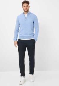 Hellblauer Strickpullover mit hohem Kragen und Reißverschluss-Detail, kombiniert mit dunkler Hose und weißen Sneakers. Glatte Textur, lässige Passform.