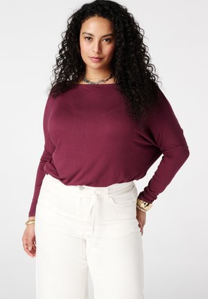 Femme aux cheveux bouclés et foncés, portant un haut bordeaux à manches longues et un pantalon blanc taille haute, posant avec les mains sur les hanches sur un fond blanc.