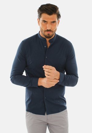 Uomo con capelli scuri e barba che indossa una camicia blu navy con bottoni e pantaloni grigio chiaro, che sistema il polsino con un'espressione seria.
