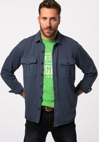 Navy-blauwe button-up shirt met twee borstzakken, gedragen over een groene graphic t-shirt. Heeft een losse pasvorm en lange mouwen.