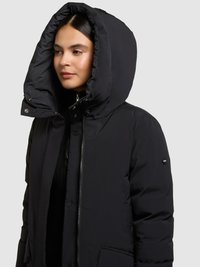 Chaqueta acolchada negra con una gran capucha, cierre de cremallera y botones, bolsillos delanteros y detalles tonales. Textura suave y diseño cálido.