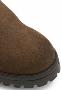 Punta de bota de ante marrón con forma redondeada, acabado texturizado y suela de goma negra con surcos profundos para un mejor agarre.