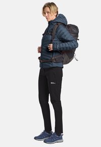 Femme portant une veste rembourrée bleue, un pantalon de randonnée noir, des chaussures de trail bleues, et portant un grand sac à dos de randonnée sombre, tournant vers la droite.