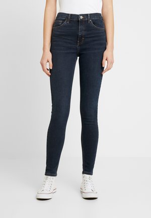 Jeans ajustados de color azul oscuro con una cintura alta, que cuentan con cinco bolsillos, costuras en contraste y un cierre de botón, combinados con zapatillas blancas.